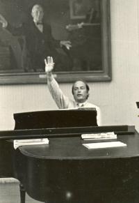 Black and white photo of Fenno Heath sitting at a piano with his right arm raised. 